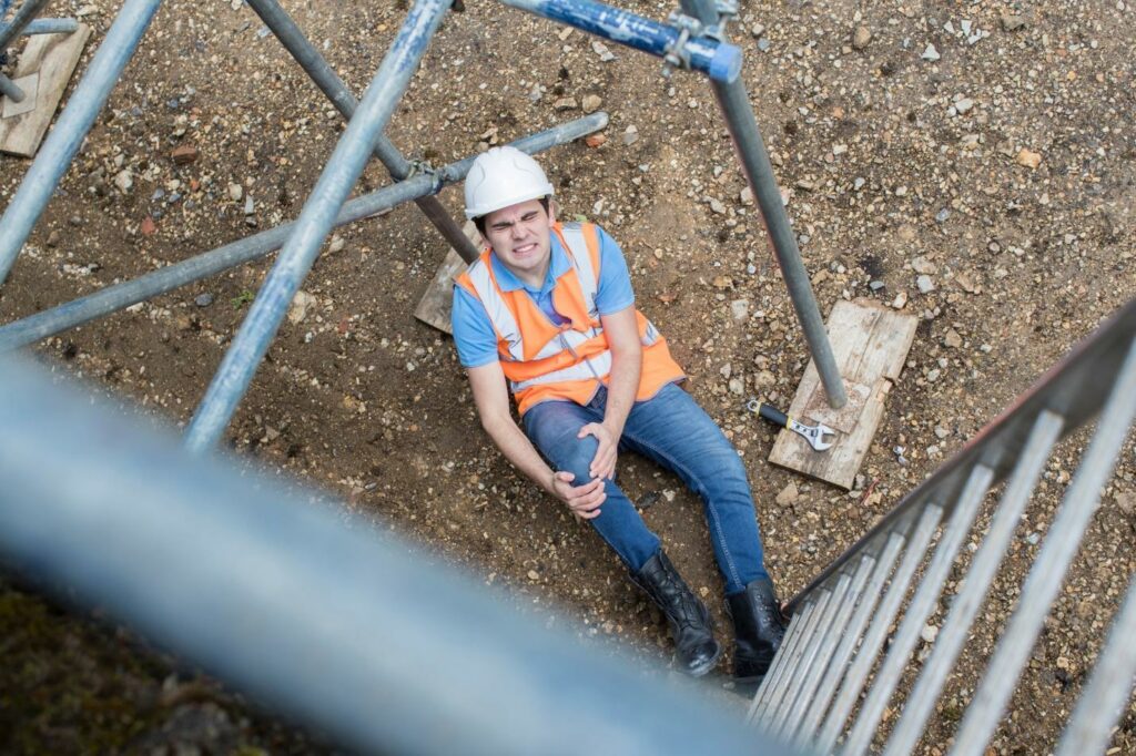 A male construction worker in a white hard hat, jeans, and orange construction vest is sitting on the ground at the base of a metal scaffolding ladder, holding his right knee with a look of pain on his face.