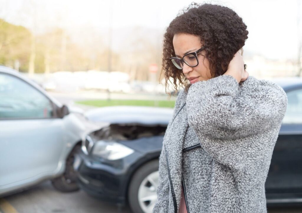 A woman rubs the back of her neck, standing in front of two crashed vehicles.