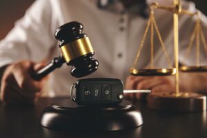 A car accident attorney holds a gavel over a car key that sits on a soundblock. A gold model of the Scales of Justice sits in the background.