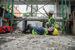 Injured worker in hard hat lying on ground in construction accident; coworker calling for help; construction site accident.