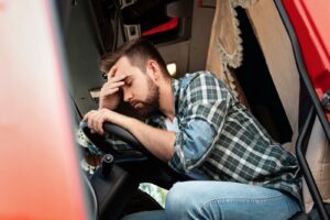 A truck driver in a plaid long sleeve shirt and jeans, sits behind the wheel of his semi-truck, his hand on his forehead, suffering from driver fatigue.