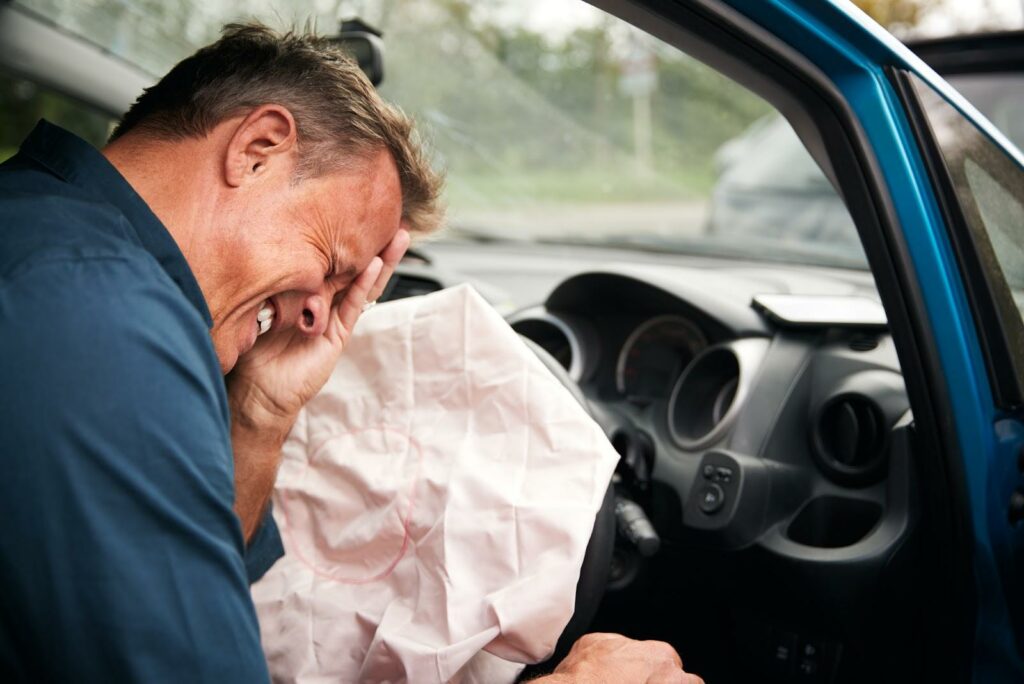 Driver holding his head in one hand, covered in bruises, seated in front of deployed airbag; airbag injuries concept.