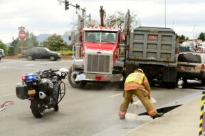 Truck accident scene in urban intersection featuring two semi trucks and an SUV in a multi-vehicle collision, with firefighter in foreground applying sand to roadway.