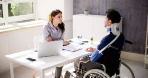 A young, dark-haired female attorney sits behind a white desk from an injured worker in a wheelchair with an arm sling and neck brace and answers his question, what does Workers’ Compensation not cover?
