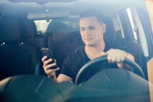 Young male driver at steering wheel looking down at mobile phone held in one hand; answer to “What is distracted driving?”