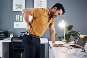 A man in dress slacks and a collared short-sleeve shirt stands over a desk, rubbing his back with a pained expression. Concept for types of workers’ compensation benefits.