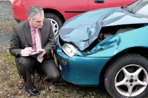 An older man dressed in a suit with a clipboard and pen examines damage to the front end of a blue-green car. Car accident insurance companies may take an adversarial approach to claims in different ways, including downplaying the damage to the vehicle.