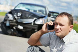 Man sitting on roadside making a call on a mobile phone next to a vehicle with a badly damaged front end; car accident lawyer concept.