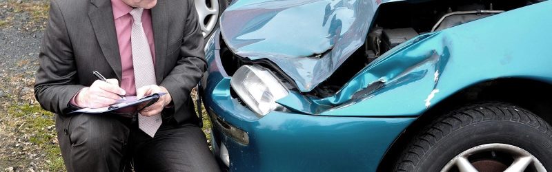 An older man dressed in a suit with a clipboard and pen examines damage to the front end of a blue-green car. Car accident insurance companies may take an adversarial approach to claims in different ways, including downplaying the damage to the vehicle.