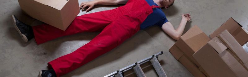 Injured employee lying on concrete floor next to fallen ladder; ladder accident at work.