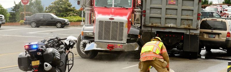 Truck accident scene in urban intersection featuring two semi trucks and an SUV in a multi-vehicle collision, with firefighter in foreground applying sand to roadway.