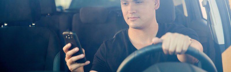 Young male driver at steering wheel looking down at mobile phone held in one hand; answer to “What is distracted driving?”