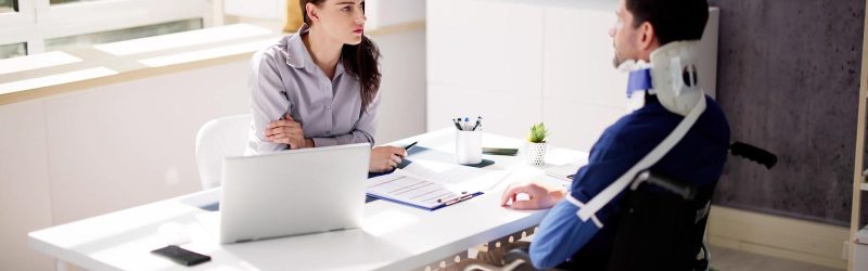 A young, dark-haired female attorney sits behind a white desk from an injured worker in a wheelchair with an arm sling and neck brace and answers his question, what does Workers’ Compensation not cover?