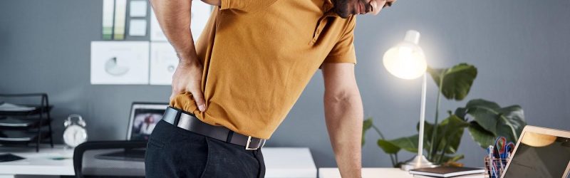 A man in dress slacks and a collared short-sleeve shirt stands over a desk, rubbing his back with a pained expression. Concept for types of workers’ compensation benefits.