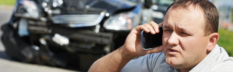 Man sitting on roadside making a call on a mobile phone next to a vehicle with a badly damaged front end; car accident lawyer concept.